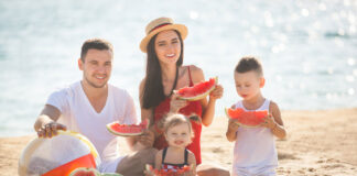 Veja opções de lanches caseiros rápidos para as crianças neste verão; família sentada na areia da praia comendo melancia