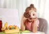 Pediatra dá dicas de como reduzir consumo de guloseimas pelas crianças; garota apoia cotovelos sobre mesa e segura um 'donut' em cada olho, na mesa há frutas e uma bebida