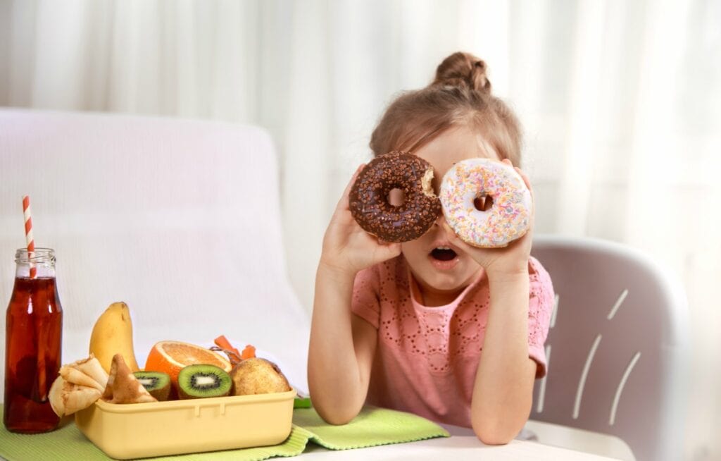 Pediatra dá dicas de como reduzir consumo de guloseimas pelas crianças; garota apoia cotovelos sobre mesa e segura um 'donut' em cada olho, na mesa há frutas e uma bebida