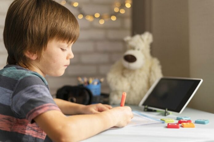 MEC autoriza atividades remotas até dezembro de 2021; garoto escreve em caderno com tela de notebook à frente