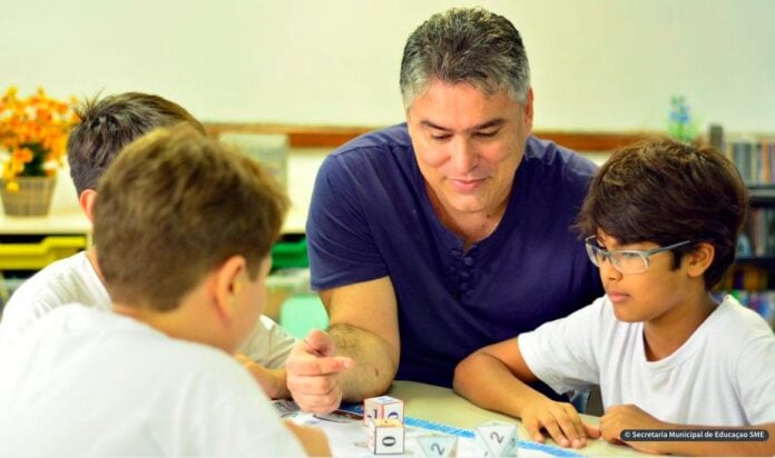Professor vence prêmio de educação científica do Rio; professor conversa com 3 alunos sobre mesa
