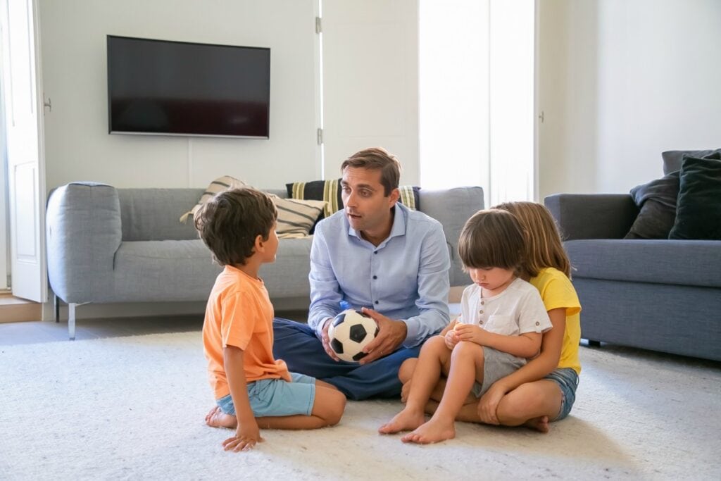 7 passos para promover a comunicação não violenta com as crianças; pai segura bola no colo e conversa com 3 crianças, todos sentados no chão