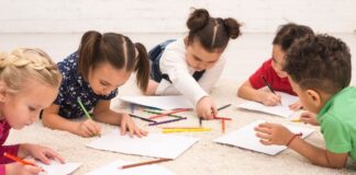 Cinco crianças deitadas no chão desenhando sobre folhas de papel com lápis de cor