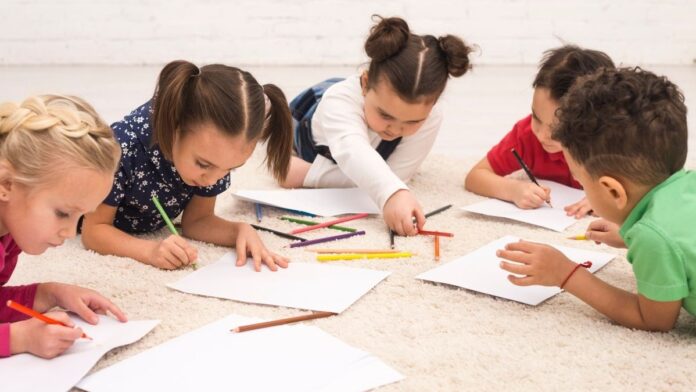 Cinco crianças deitadas no chão desenhando sobre folhas de papel com lápis de cor