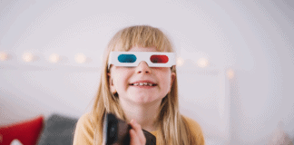 Quanto maior o tempo de TV da criança, maior o estresse dos pais; menina loira com óculos 3D sorri e segura controle remoto com a mão