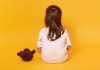 Projeto busca conscientizar crianças sobre abuso sexual; criança de cabelo liso grande está sentada no chão de costa com urso de pelúcia ao lado em imagem de fundo amarelo