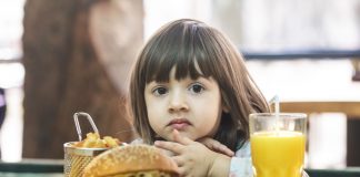 Gordura e açúcar na infância podem alterar o microbioma por toda a vida; criança pequena segura batata frita que está sob mesa junto a hambúrguer e suco