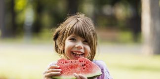 9 alimentos que ajudam na hidratação das crianças; menina sorridente de macacão jeans e blusa rosa segura melancia com as duas mãos