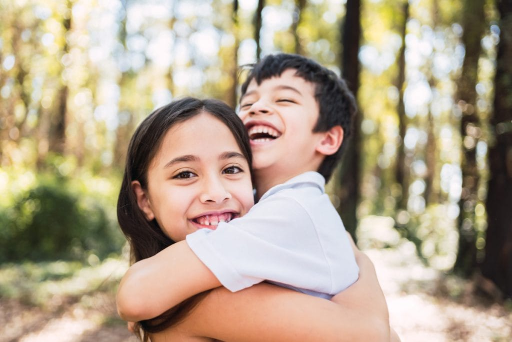 Cada filho é único: saiba como agir para despertar o amor entre os irmãos; garota abraça irmão mais novo sorridente em espaço aberto com árvores ao fundo