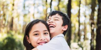 Cada filho é único: saiba como agir para despertar o amor entre os irmãos; garota abraça irmão mais novo sorridente em espaço aberto com árvores ao fundo