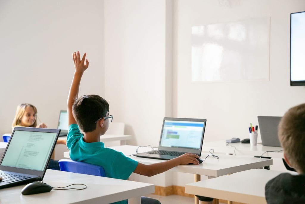 A tecnologia deve servir à vida e resgatar a alma das crianças na escola; aluno sentado à frente de mesa com computador levanta o braço em sala de aula