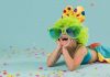 Fantasias de Carnaval para as crianças aproveitarem a folia em casa; menina está deitada no chão com rosto apoiado nas mãos. Ela veste fantasia colorida, peruca verde-limão e óculos de plástico verde.