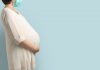 Grávidas repassam anticorpos da Covid-19 aos bebês, indica estudo; mulher grávida de vestido branco e cabelo curto estende braço por baixo da barriga e usa máscara