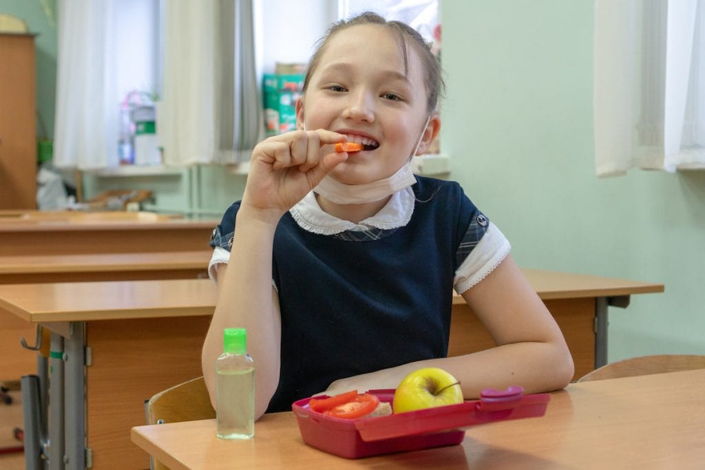 Lanches saudáveis para levar na escola; Menina asiática comendo um pedaço de cenoura. A frente dela, uma lancheira com frutas e um vidro de alcool em gel sobre a mesa.
