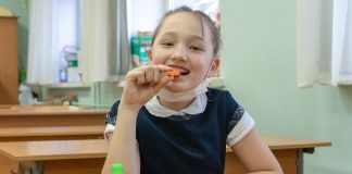 Lanches saudáveis para levar na escola; Menina asiática comendo um pedaço de cenoura. A frente dela, uma lancheira com frutas e um vidro de alcool em gel sobre a mesa.