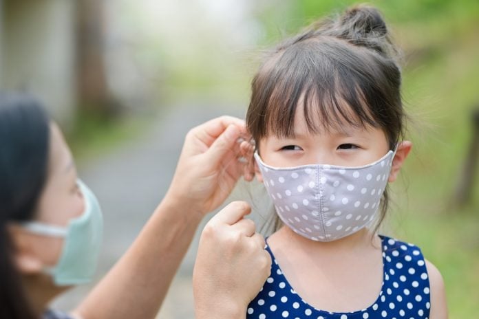 Máscara para crianças: qual é a mais segura agora?; mulher adulta mexe na máscara de tecido de cor cinza com bolinhas que menina oriental está usando