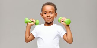 Musculação na infância: conheça quais são os riscos e os benefícios da prática; Menino segura dois halteres verdes na altura dos ombros