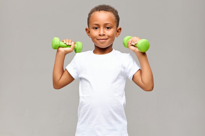 Musculação na infância: conheça quais são os riscos e os benefícios da prática; Menino segura dois halteres verdes na altura dos ombros