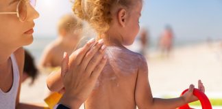 Saiba como usar protetor solar e repelente nas crianças; mão de adulto passa protetor solar nas costas de criança