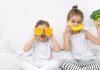 10 alimentos para garantir uma boa noite de sono para os pequenos! Duas meninas sentadas em uma cama branca. A menor segura duas metades de laranja na altura dos olhos. A maior segura uma banana na altura da boca, simulando um sorriso e um brócolis na outra.