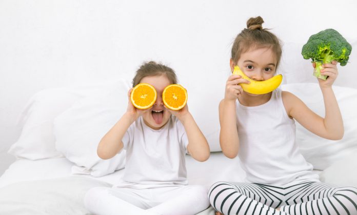 10 alimentos para garantir uma boa noite de sono para os pequenos! Duas meninas sentadas em uma cama branca. A menor segura duas metades de laranja na altura dos olhos. A maior segura uma banana na altura da boca, simulando um sorriso e um brócolis na outra.