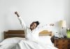 Xô, preguiça! Um exercício mental para levantar da cama com ânimo; mulher negra de camisa branca se espreguiça na cama sorridente com os braços abertos