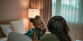 Qual a diferença entre amar e mimar os filhos?; mãe negra de costas olha para filha negra que ri e está com cotovelos apoiados sobre sofá