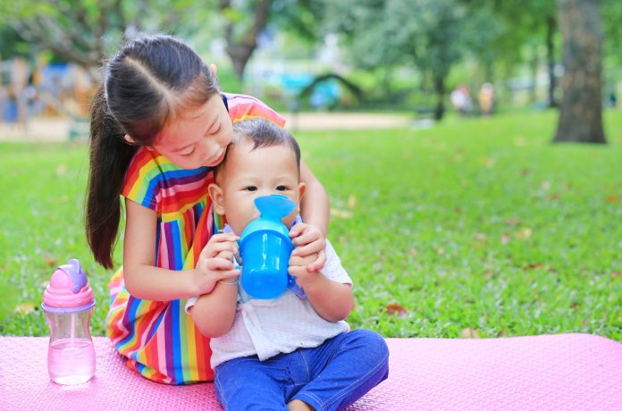 Leite de vaca, fórmulas e compostos lácteos: saiba quando são indicados; bebê asiático sentado sobre grama no parque segura copo de transição com ajuda da irmã mais velha