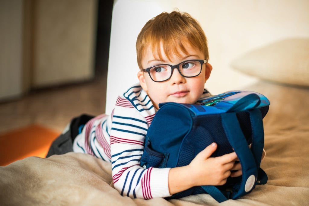 2020: o ano que mais prejudicou a visão das crianças; menino de óculos apoia tronco sobre cama e segura bolsa com os dois braços