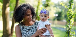 Guia traz 12 passos para a alimentação saudável de bebês; mãe de cabelos pretos segura bebê nos braços em parque ao ar livre