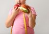 Estudo indica a possível causa da obesidade na infância + Menina segurando um lanche em uma mão e na outra uma maçã que está na direção da boca. Ela usa camiseta rosa e em seus ombros tem uma fita métrica.