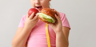Estudo indica a possível causa da obesidade na infância + Menina segurando um lanche em uma mão e na outra uma maçã que está na direção da boca. Ela usa camiseta rosa e em seus ombros tem uma fita métrica.