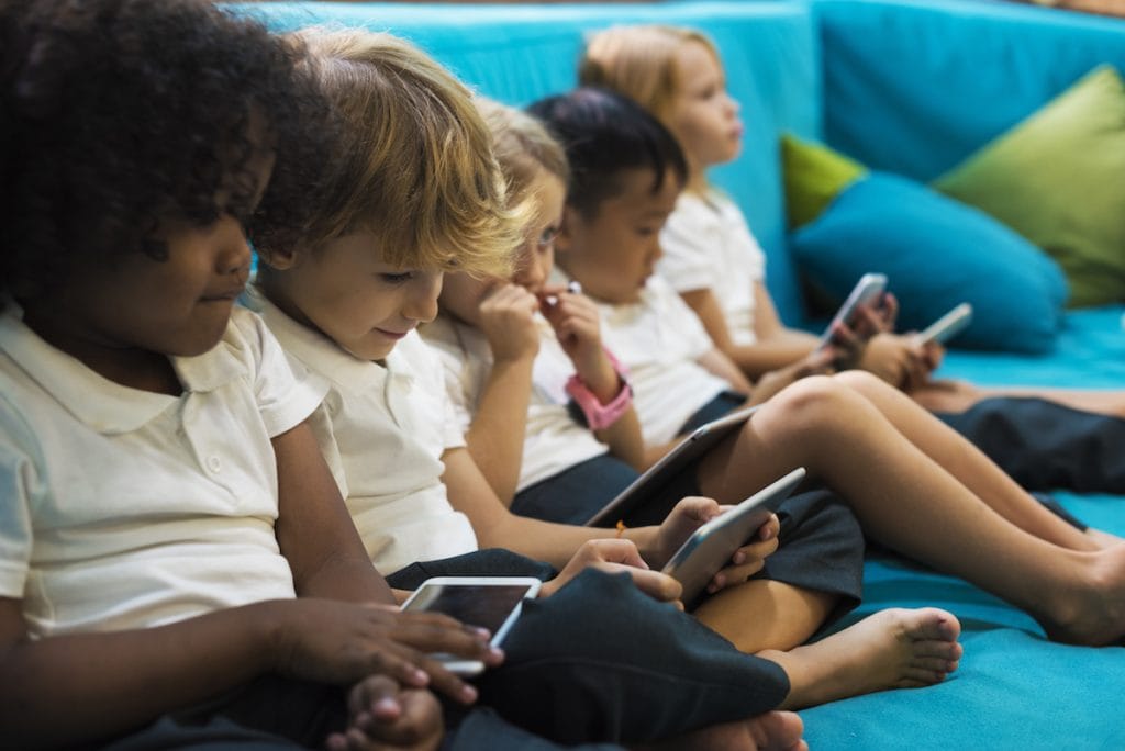 Tecnologia aliada à educação: crianças sentadas em sofá azul manuseiam smartphones ou tablets, cada uma, com o seu