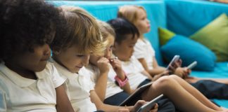 Tecnologia aliada à educação: crianças sentadas em sofá azul manuseiam smartphones ou tablets, cada uma, com o seu