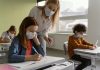 Piora da pandemia não tem relação com as escolas abertas; professora de máscara em pé fala com aluna de máscara sentada em carteira escolar