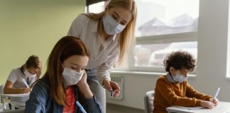 Piora da pandemia não tem relação com as escolas abertas; professora de máscara em pé fala com aluna de máscara sentada em carteira escolar