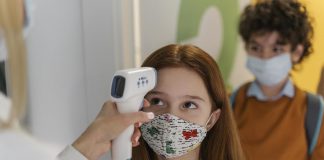 Reabertura das escolas particulares: 13 estados e o Distrito Federal retomam aulas presenciais; mão de adulto segura termômetro para aferir temperatura de menina de máscara