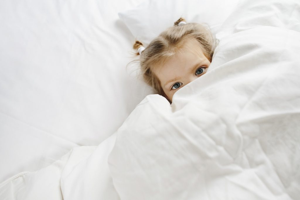 Masturbação infantil: como conversar sobre isso com os filhos; menina de olhos claros está deitada em cama coberta com lençol branco até os olhos