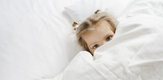 Masturbação infantil: como conversar sobre isso com os filhos; menina de olhos claros está deitada em cama coberta com lençol branco até os olhos