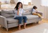 6 práticas comuns de alienação parental na pandemia; mãe sentada no sofá fala ao telefone e tem braço esticado para o lado com palma da mão para cima, n canto do sofá criança está sentada encolhida