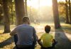 O silêncio, como resposta, pode ser útil para o filho; pai e filho estão em área verde, com árvores, sentados no chão de costas para a câmera