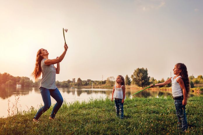 Muitos pais têm medo de errar com os filhos, mas isso faz parte da jornada; mulher rebate peteca com raquete e duas crianças olham para ela em área verde ao ar livre