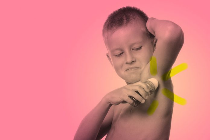 Crianças com mau cheiro nas axilas podem usar desodorante infantil?; menino sem camisa passa desodorante na axila, em imagem de fundo rosa