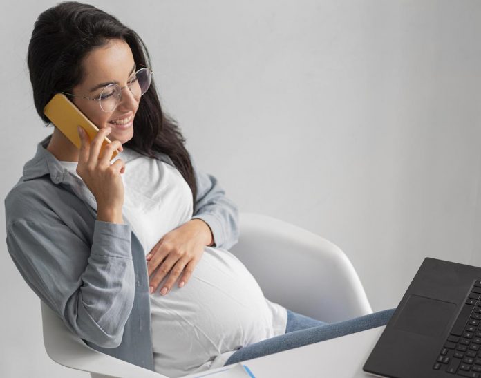 Agora é lei: grávidas devem realizar trabalho remoto; gestante de óculos conversa ao telefone com notebook à frente