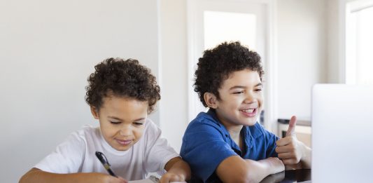 Em plena pandemia, é uma boa ideia mudar de escola?; dois irmãos sorridentes estudam – um escreve no caderno e outro olha para a tela de notebook