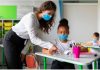 Afinal, quem cuida dos professores na pandemia?; professora de máscara está em pé e aponta para folha de aluna de máscara sentada