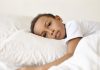 Enurese noturna: saiba como prevenir o xixi na cama durante o sono em crianças; menino moreno está deitado na cama com cabeça sobre travesseiro branco e olhos abertos com expressão séria