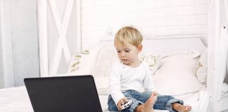 Tecnologia na infância: prejuízos que o excesso das telas pode trazer para as crianças; bebê sentado na cama olhando para computador online