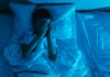 Os 4 distúrbios do sono que se agravaram na pandemia; menino deitado na cama em quarto escuro coloca mãos sobre rosto