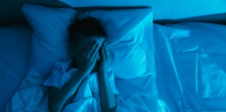 Os 4 distúrbios do sono que se agravaram na pandemia; menino deitado na cama em quarto escuro coloca mãos sobre rosto